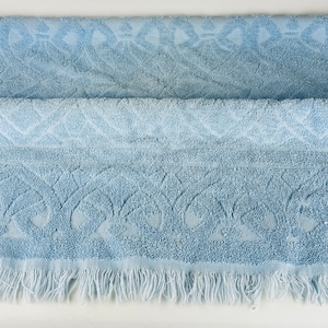 May include: A pale blue bath towel with a decorative pattern and a fringed edge. The towel is folded, revealing the detailed design. The pattern features stylized floral and geometric shapes.