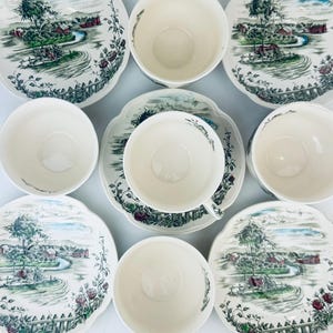 Puede incluir: Vista cenital de un juego de té color crema con seis tazas y platillos. Cada platillo presenta un diseño de paisaje escénico con un río, árboles y una casa. Las tazas tienen un diseño simple y elegante.
