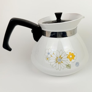 Op de afbeelding: Witte vintage koffiepot met een zwarte handgreep en deksel. De pot heeft een bloemenontwerp in geel, blauw en groen. Een zilveren band omcirkelt het midden van de pot. De pot is waarschijnlijk gemaakt van glas of keramiek.