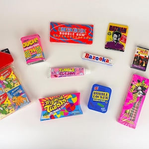 Puede incluir: Una colección de paquetes y recipientes de chicle vintage, incluyendo diseños como "Dino Bones", "Checkbook", "Bazooka" y "Batman". El surtido colorido presenta varias formas y tamaños, con embalajes en rosa brillante, azul y amarillo.