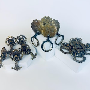 Puede incluir: Un surtido de herrajes de gabinete de latón antiguo, que incluyen tiradores y perillas ornamentadas. Los herrajes presentan intrincados diseños florales y de volutas, con una estética vintage. Las piezas están dispuestas sobre pedestales blancos.