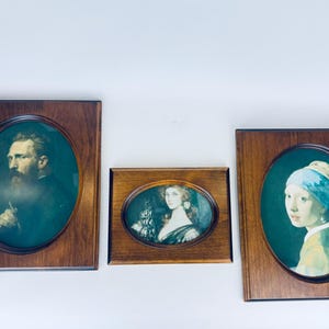 May include: Three framed oval portraits with dark wood frames. The portraits depict classic art pieces, including a man with a beard, a woman in a dress, and a woman with a blue headscarf. The frames are rectangular.