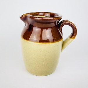 May include: A ceramic pitcher featuring a two-tone design with a glossy dark brown glaze on the upper portion and a speckled cream-colored glaze on the lower body. The pitcher includes a curved handle and a small pouring spout at the rim.