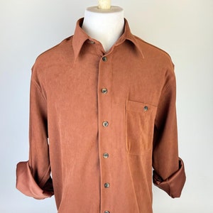 May include: A button-down shirt in a reddish-brown colour, with long sleeves and a classic collar. The shirt has a chest pocket and buttons down the front. The sleeves are cuffed, showing a darker, satin-like lining.
