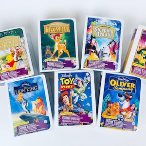 Op de afbeelding: Een verzameling van zeven Disney VHS-banden met klassieke animatiefilms. Titels zijn onder meer "Winnie the Pooh", "Bambi", "Sleeping Beauty", "The Three Caballeros", "The Lion King", "Toy Story" en "Oliver & Company". Elke band heeft kleurrijke omslagkunst.
