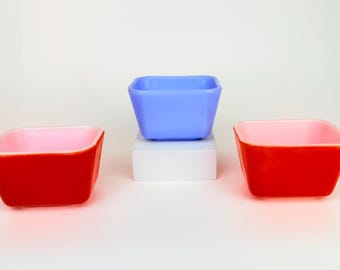 May include: Three square-shaped bowls in the image. Two are red with pink interiors, and one is light blue. The blue bowl sits on a white block. The bowls are empty and have a retro aesthetic.