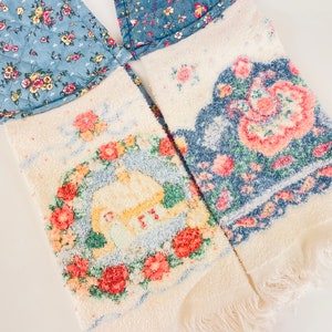 Op de afbeelding: Twee witte keukendoeken met bloemenpatronen. Een handdoek heeft een blauw bloemenpatroon met een witte cottage-scène. De andere handdoek heeft een blauw en roze bloemenpatroon.