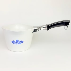 Op de afbeelding: Witte steelpan met een zwarte handgreep en een blauw bloemenontwerp. De steelpan heeft een zilveren metalen bevestiging voor de handgreep en een kleine lus aan het uiteinde. Het bloemenontwerp bevindt zich aan de voorkant van de steelpan.