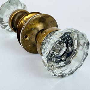 May include: A set of antique door handles, featuring two clear, faceted glass knobs with brass-toned metal parts. The knobs have a vintage aesthetic, showcasing a classic design. The hardware is likely for a door.