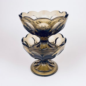 Puede incluir: Un plato de servir de dos niveles, de estilo vintage, en un tono gris-marrón ahumado. El plato de cristal presenta un borde festoneado y un diseño floral. La base es un pedestal, y los dos cuencos están conectados por un tallo central.