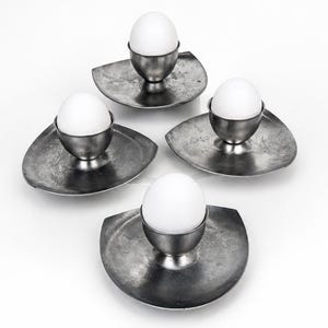 May include: A set of four silver-toned metal egg cups, each featuring a smooth, rounded cup centred on a wide, flat, irregularly shaped base. Each cup holds a single white egg, creating a uniform arrangement of four individual serving pieces.