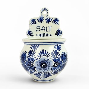 Può includere: Un salino da parete in ceramica bianca con motivi floreali dipinti a mano in blu e la parola SALT stampata in lettere nere sul pannello posteriore superiore. Il contenitore ha un corpo arrotondato e un foro superiore per appenderlo.