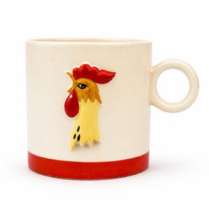 Puede incluir: Una taza de cerámica de color crema con un diseño en relieve de una cabeza de gallo en la parte frontal. El gallo tiene una cresta y barbillas de color rojo brillante con plumas amarillas y marrones. La taza tiene un asa circular y una base roja.