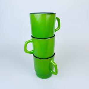 May include: Three green ceramic mugs stacked on top of each other. The mugs have a black rim and a glossy finish.