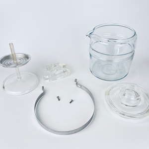 May include: Disassembled parts of a glass coffee maker, including the carafe, lid, filter basket, and metal band.