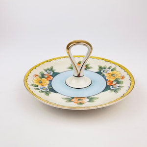 Könnte beinhalten: Ein Vintage-Serviertablett aus Keramik mit Griff. Das Tablett hat eine hellblaue Mitte, ein Blumendesign mit gelben, orangefarbenen und blauen Blumen und einen goldfarbenen Griff. Der Rand hat einen gewellten Rand.