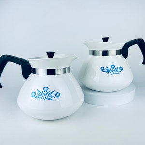May include: Two white coffee pots with black handles and lids. Each pot has a silver band around the middle and a blue floral design. The pots are on a white surface.