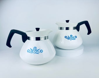 Op de afbeelding: Twee witte koffiepotten met zwarte handvatten en deksels. Elke pot heeft een zilveren band en een blauw bloemenontwerp. De potten staan op een wit oppervlak.