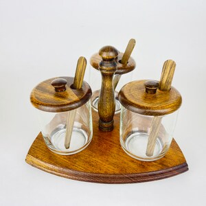 Puede incluir: Un estante de especias de madera con tres tarros de vidrio y tapas de madera. Cada tarro tiene una cuchara de madera en su interior. El estante está hecho de madera oscura y tiene una forma redondeada.