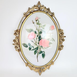 May include: An oval wall mirror featuring a gold-toned metal frame with ornate leaf and rose bud detailing. The center glass displays a botanical illustration of pink roses with green leaves and stems against a white background.