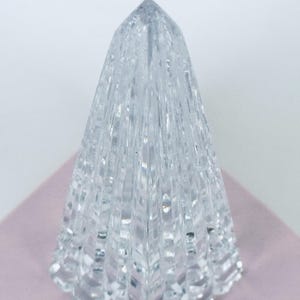 May include: A clear, faceted crystal obelisk with a pointed top. The crystal is placed on a light pink surface, and the background is white. The obelisk has a textured, geometric design.