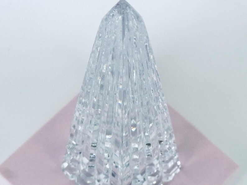 May include: A clear, faceted crystal obelisk with a pointed top. The crystal is placed on a light pink surface, and the background is white. The obelisk has a textured, geometric design.