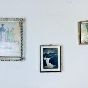 May include: Three framed vintage art pieces hanging on a white wall. The largest frame is silver with a detailed border, showcasing a watercolor of a fountain. The middle frame is silver with a black and white landscape. The smallest frame is gold with a floral print.