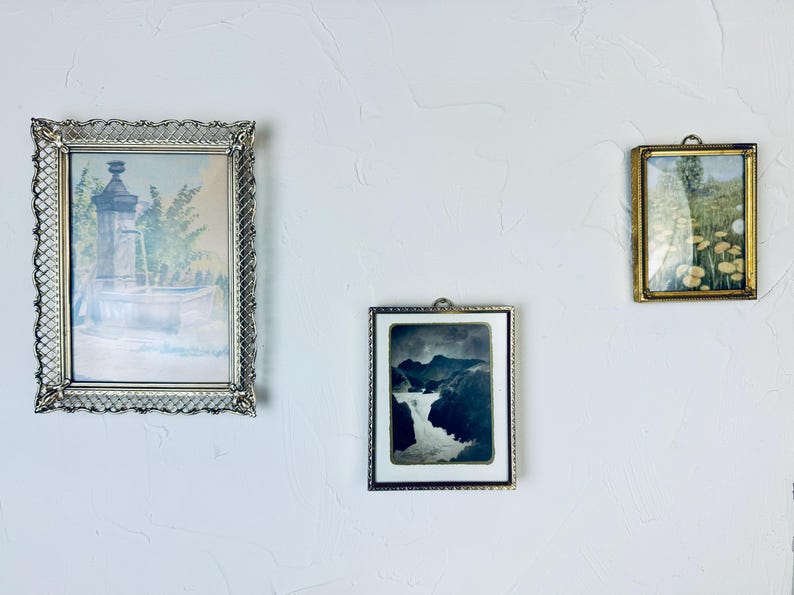 May include: Three framed vintage art pieces hanging on a white wall. The largest frame is silver with a detailed border, showcasing a watercolor of a fountain. The middle frame is silver with a black and white landscape. The smallest frame is gold with a floral print.
