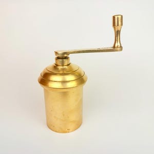 Può includere: Un antico utensile da cucina in ottone con un corpo cilindrico e una manovella. L'utensile ha una finitura dorata ed è progettato per la preparazione di cibi. La manovella è attaccata alla parte superiore del cilindro e il manico è verticale. L'utensile viene probabilmente utilizzato per macinare o pressare.