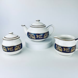 May include: A white ceramic tea set with gold accents and a blue and gold band featuring the text "Tetley Teas". The set includes a teapot, creamer, and sugar bowl.