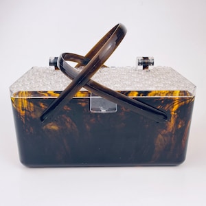 May include: A vintage-style, rectangular handbag with a tortoiseshell pattern. The clear lid is textured with small, clear, rounded bumps. The handle is a dark brown color, and the bag has a clear clasp. The bag is made of a hard plastic material.
