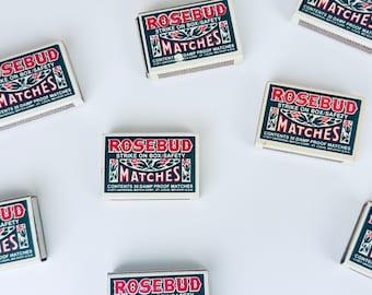 以下が含まれることがあります： 白い背景に、赤と緑で「ROSEBUD MATCHES」と書かれたマッチ箱がいくつか配置されています。各箱には内容と安全に関する情報が記載されており、ヴィンテージスタイルです。