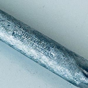 May include: Close-up of a silver metal rod with a textured surface and markings. The rod is about 10 inches long.