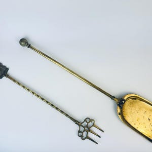 May include: Two antique brass fireplace tools. One is a long-handled shovel with a rectangular scoop. The other is a poker with a decorative handle and a three-pronged fork. The tools are golden in colour and appear to be vintage.