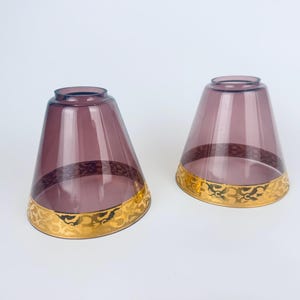 May include: Two vintage purple glass shades with a gold filigree design around the base. The shades have a flared shape and are likely for a lamp or chandelier. The gold trim adds a touch of elegance.