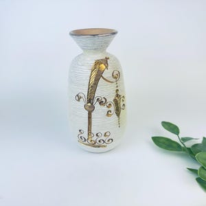 May include: White ceramic vase with gold leaf design. The vase has a textured surface and a narrow neck.
