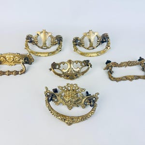 May include: Collection of six antique brass drawer pulls in various ornate designs. The pulls feature curved handles and decorative backplates with floral and scrollwork details. The brass has a warm, aged patina.