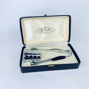 Könnte beinhalten: Ein Vintage-Bar-Tool-Set in einer schwarzen Box mit cremefarbenem Satinfutter. Das Set enthält einen Chrom-Eiskübel, Eiszangen und einen Barlöffel mit dunklem Holzgriff. Der Deckel der Box trägt den Text "Barmates Gold".