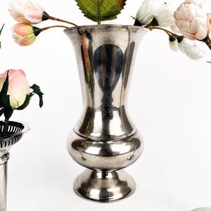 May include: Three silver-toned metal vases of varying shapes and sizes. The center vase has a bulbous base and flared rim. The left vase features a decorative pierced rim, and the right vase is tall and slender. Artificial pink and white flowers.