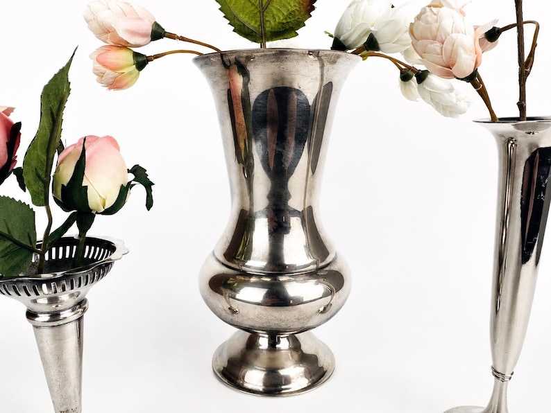 May include: Three silver-toned metal vases of varying shapes and sizes. The center vase has a bulbous base and flared rim. The left vase features a decorative pierced rim, and the right vase is tall and slender. Artificial pink and white flowers.