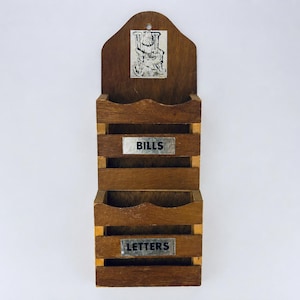 May include: A wooden wall-mounted mail organiser with two compartments. The top section has a decorative cutout, and the bottom sections are labelled "BILLS" and "LETTERS" on metal plates. The wood has a warm, brown tone.