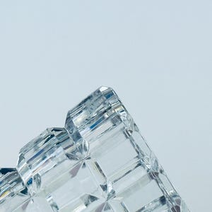 May include: Close-up of a clear, faceted crystal object. The object has a geometric design with multiple rectangular and angled surfaces. The crystal reflects light, creating a sparkling effect against a light blue background.