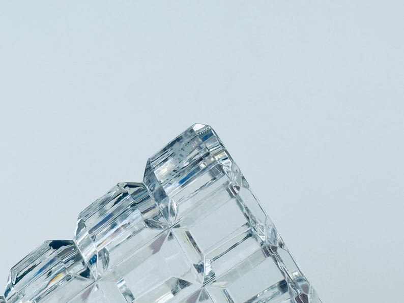 May include: Close-up of a clear, faceted crystal object. The object has a geometric design with multiple rectangular and angled surfaces. The crystal reflects light, creating a sparkling effect against a light blue background.