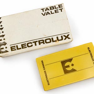 Puede incluir: Un valet de mesa rectangular amarillo Electrolux Canada con logotipo y texto en negro, acompañado de su caja de cartón blanca original que presenta el nombre de la marca y una serie de iconos gráficos circulares en el lado izquierdo del envase.