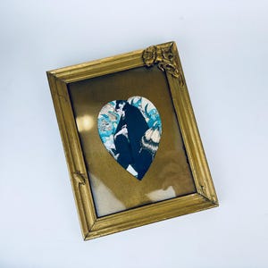 Puede incluir: Un marco de fotos dorado con una abertura en forma de corazón. La abertura contiene una impresión vintage de una mujer con un vestido negro con un diseño blanco y azul.