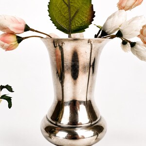 May include: A polished silver-toned metal vase with a flared rim and a rounded base, containing several artificial flowers with pale pink and white petals and green leaves, alongside a small silver-toned bowl with a scalloped edge.