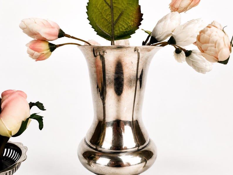 May include: A polished silver-toned metal vase with a flared rim and a rounded base, containing several artificial flowers with pale pink and white petals and green leaves, alongside a small silver-toned bowl with a scalloped edge.