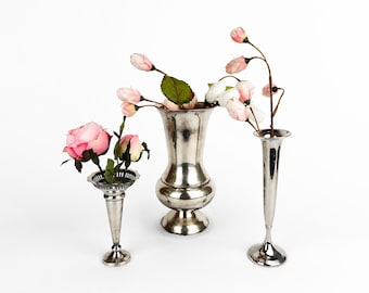 May include: Three silver vases of varying shapes, each holding artificial flowers. The flowers are primarily pink and white, with green leaves. The vases are arranged against a white background.
