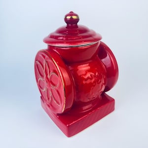 Op de afbeelding: Rode keramische pot met een deksel en een bloemenmotief. De pot heeft een vierkante basis en een rond lichaam. Het deksel heeft een gouden rand.