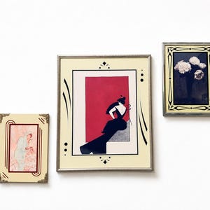 May include: Three framed wall art pieces. The left frame shows a light-coloured figure in a pink setting. The centre frame features a black-clad figure against a red rectangle. The right frame displays a bouquet of white flowers on a dark background.